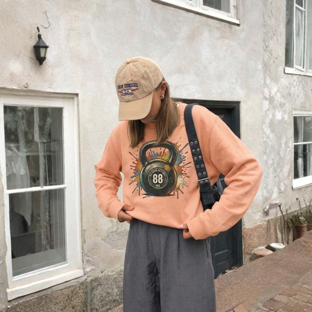 Person wearing a peach sweatshirt with a kettlebell design, standing outside a building.