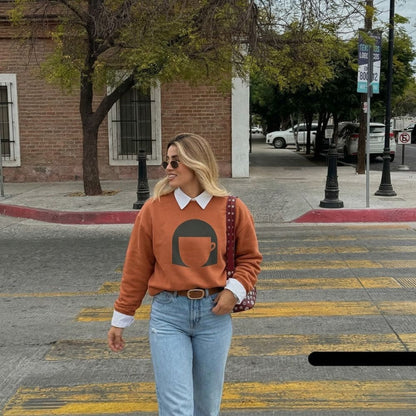 Woman walking on a street wearing a brown sweater with a coffee cup design, light blue jeans, and sunglasses.