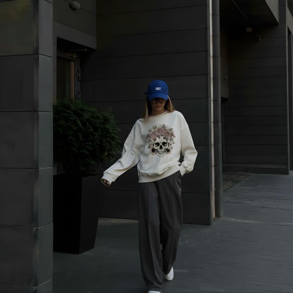 Person wearing a white sweatshirt with a skull design and a blue cap, standing in front of a dark building.