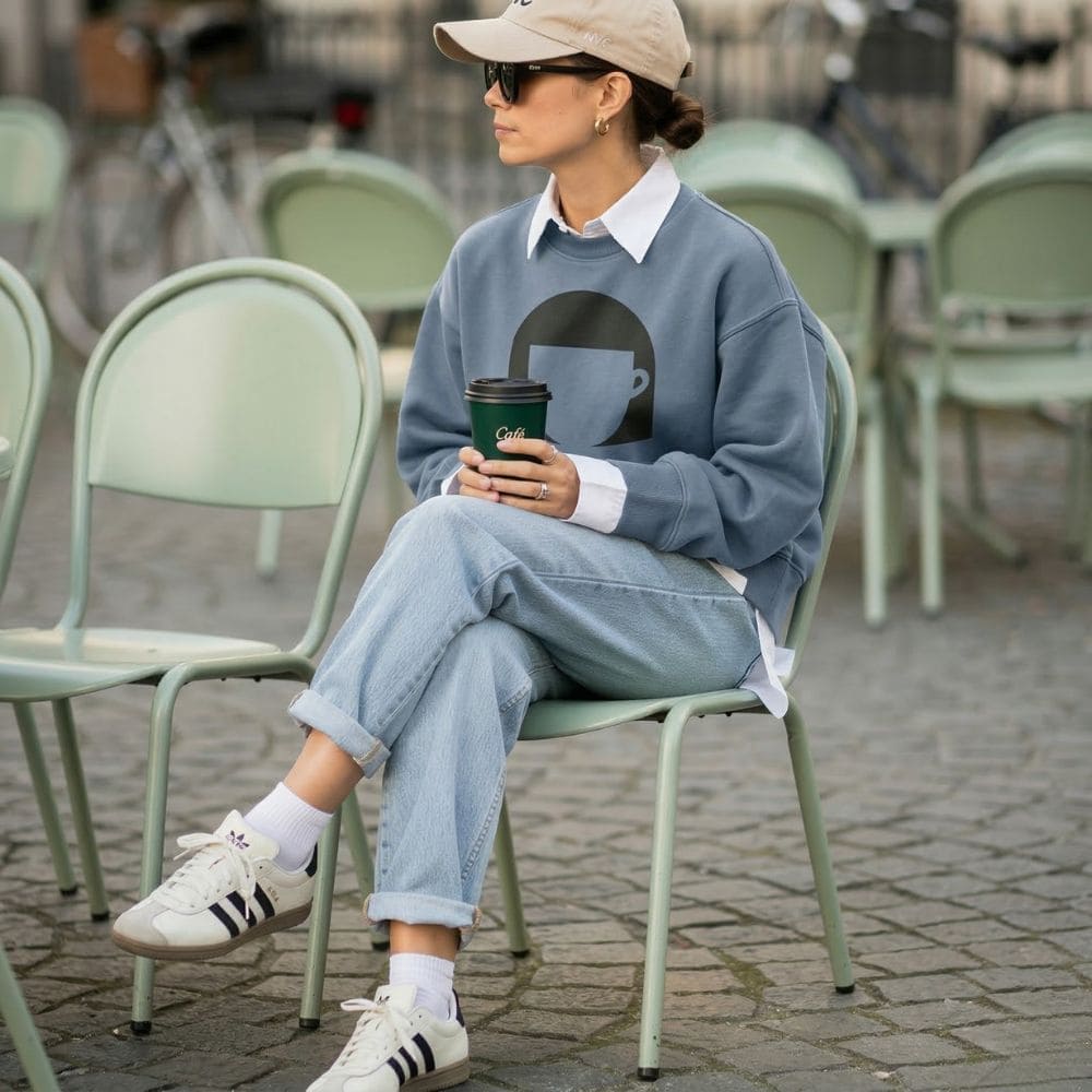 Person sitting on a chair outdoors, wearing a blue sweatshirt with a coffee cup design, light blue jeans, and Adidas sneakers.