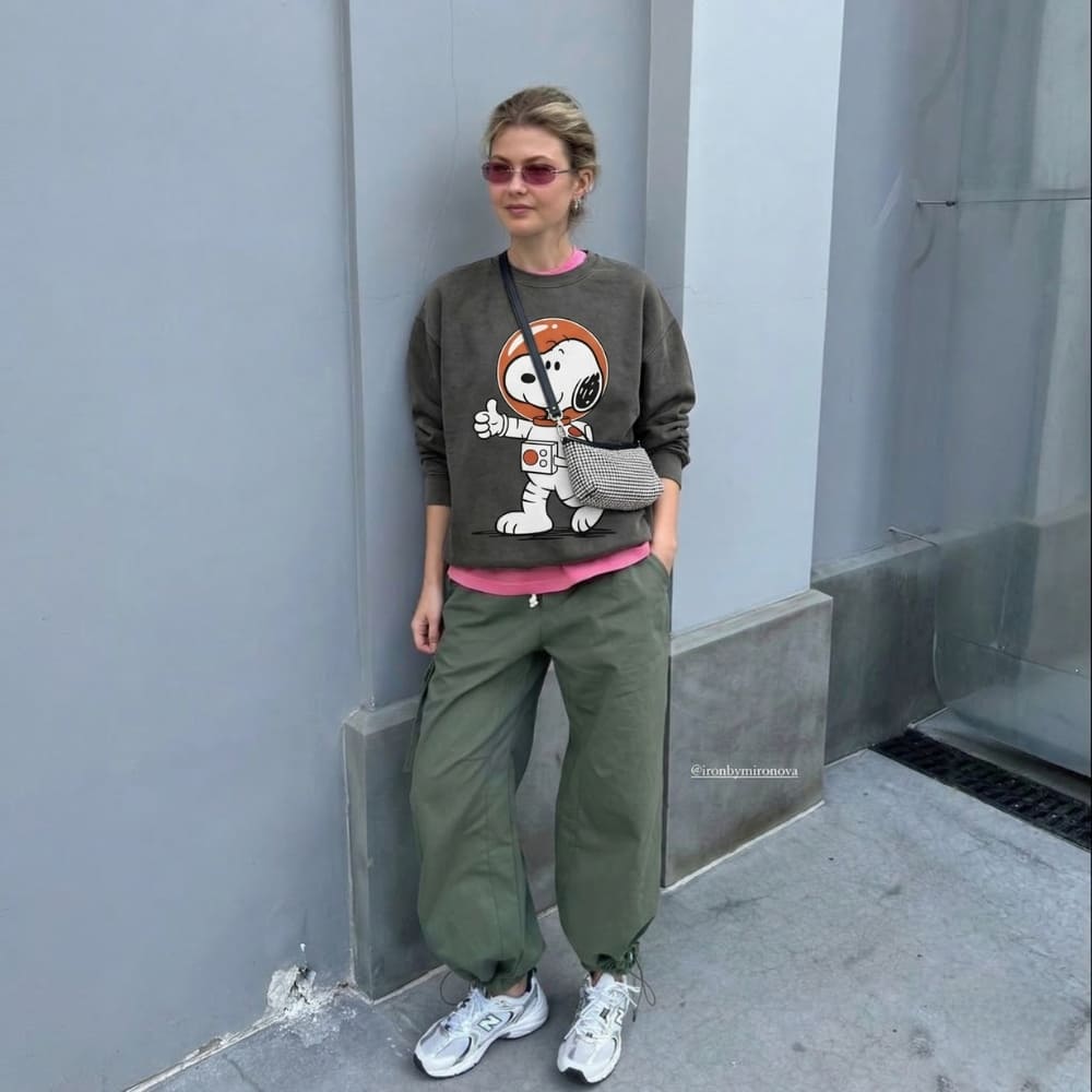 Person wearing a graphic sweatshirt and green pants standing against a gray wall.
