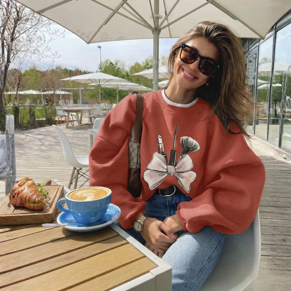 Woman in a red sweatshirt with a bow and lipstick design, sitting at an outdoor cafe table with a cup of coffee and pastry.