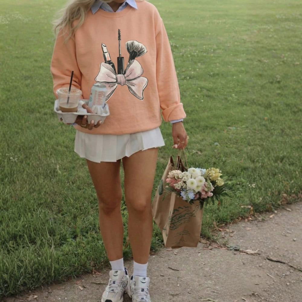 Person wearing a peach sweatshirt with a bow design, holding a drink and flowers on a grassy path.