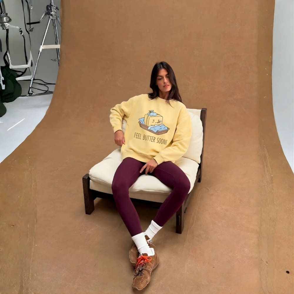 Person wearing a yellow sweater with a graphic design, sitting on a chair against a brown backdrop.