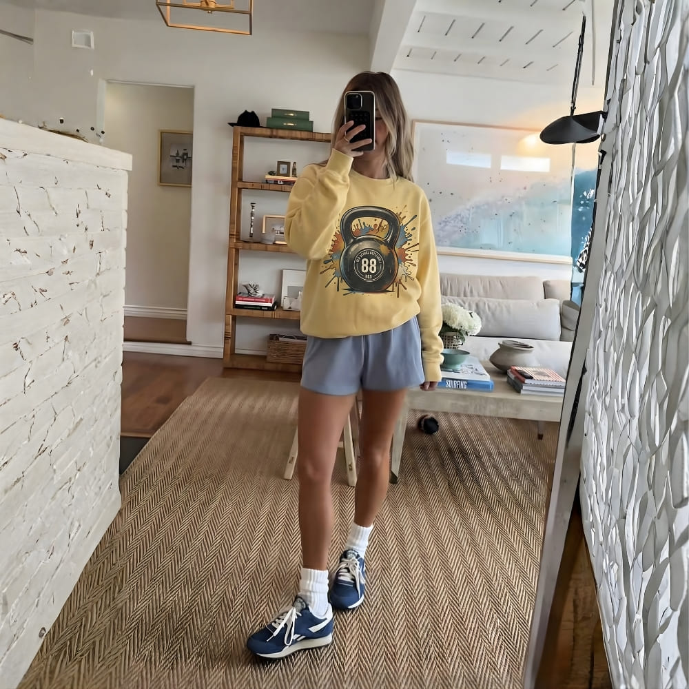 Person taking a mirror selfie wearing a yellow sweatshirt with a kettlebell design, blue shorts, and sneakers in a living room.