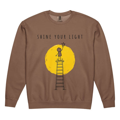 Brown sweatshirt with a graphic of a person on a ladder reaching for a star, set against a yellow circle with 'Shine Your Light' text.