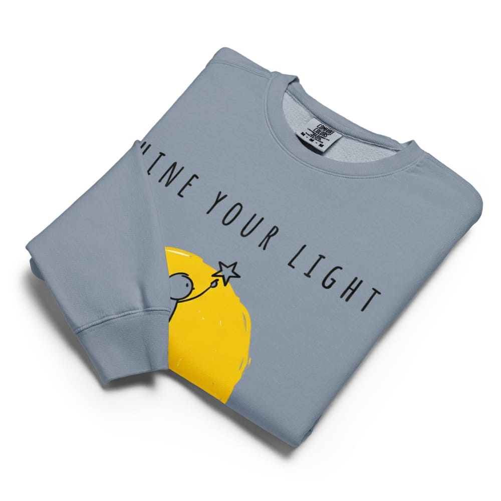 Folded blue sweatshirt with yellow graphic and text 'Shine Your Light' on a white background