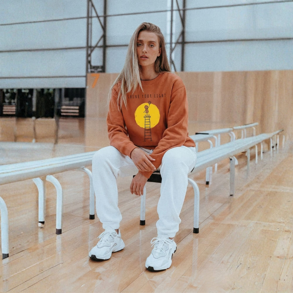 Person wearing a orange sweatshirt with a yellow graphic sitting on a bench indoors.