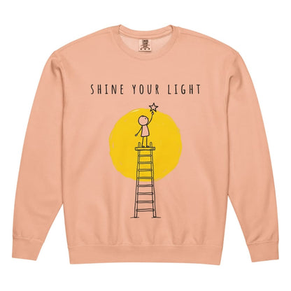 Peach-colored sweatshirt with 'Shine Your Light' graphic and text on a white background