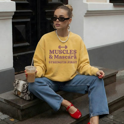 Woman wearing a yellow sweatshirt with 'MUSCLES & Mascara STRENGTH FIRST' text, sitting on steps.