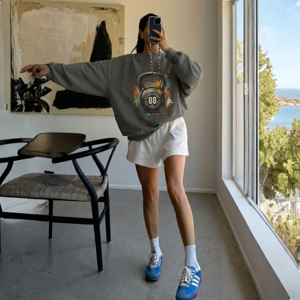 Person taking a mirror selfie wearing a gray sweatshirt with a kettlebell design, white shorts, and blue sneakers in a room with large windows.
