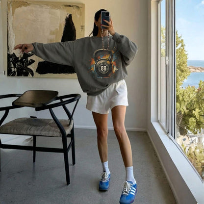 Person taking a mirror selfie wearing a gray sweatshirt with a kettlebell design, white shorts, and blue sneakers in a room with large windows.