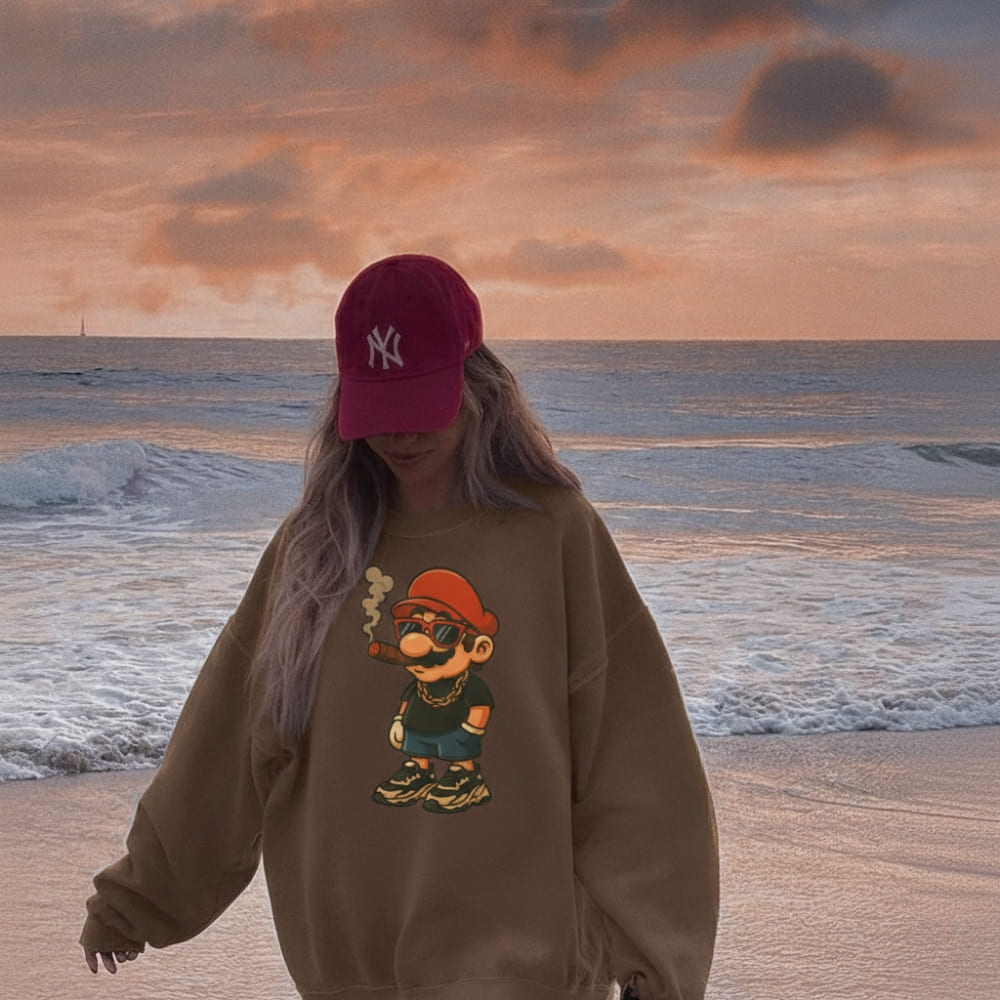 Person wearing a brown sweatshirt with a graphic design and a red cap on a beach at sunset.