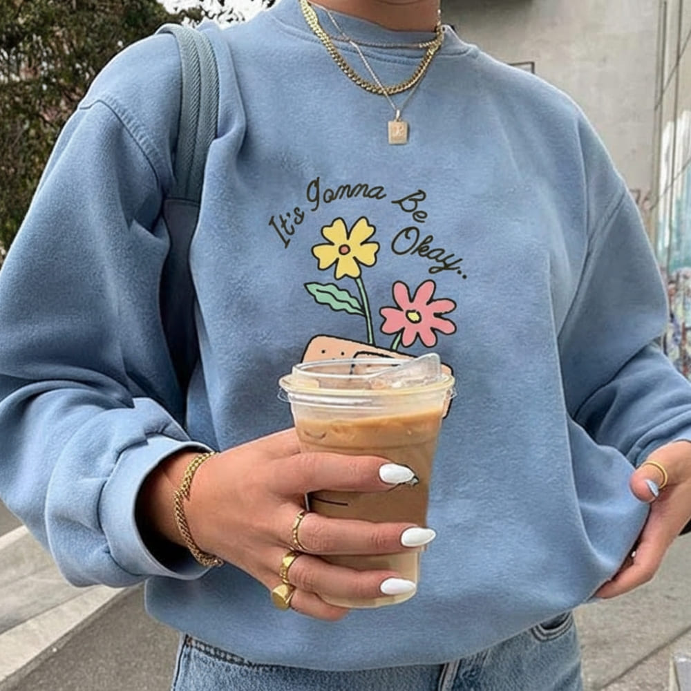 Person wearing a blue sweatshirt with a motivational quote and flowers, holding an iced coffee.