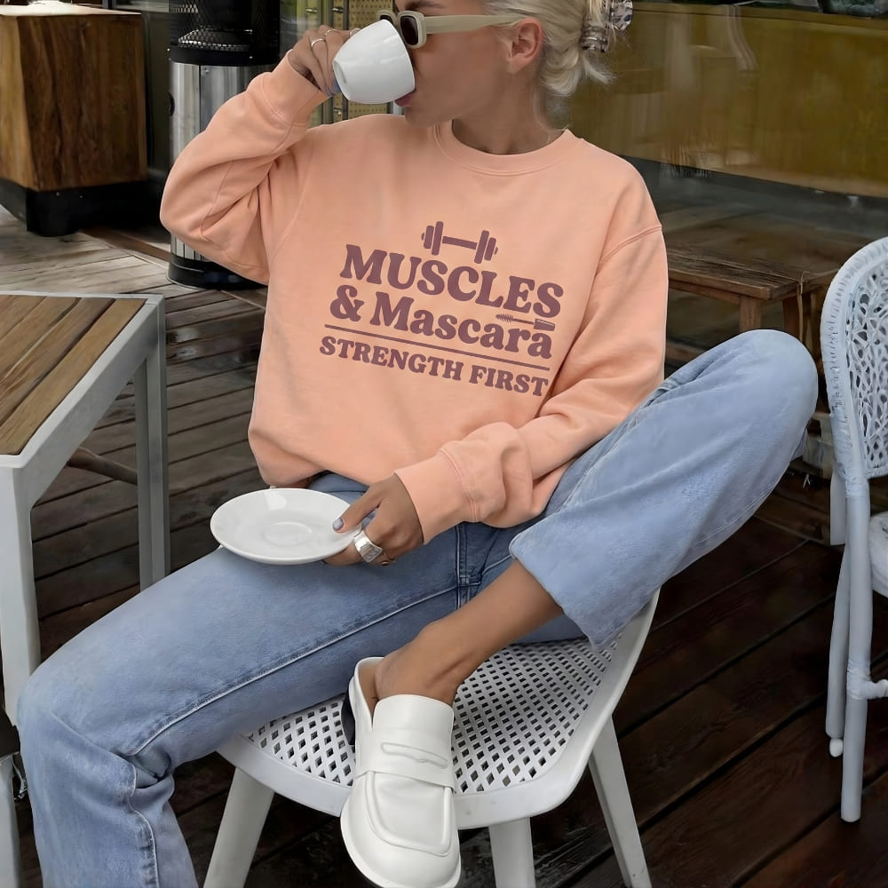 Person wearing a peach-colored sweatshirt with 'Muscles & Mascara Strength First' text, sitting outdoors.