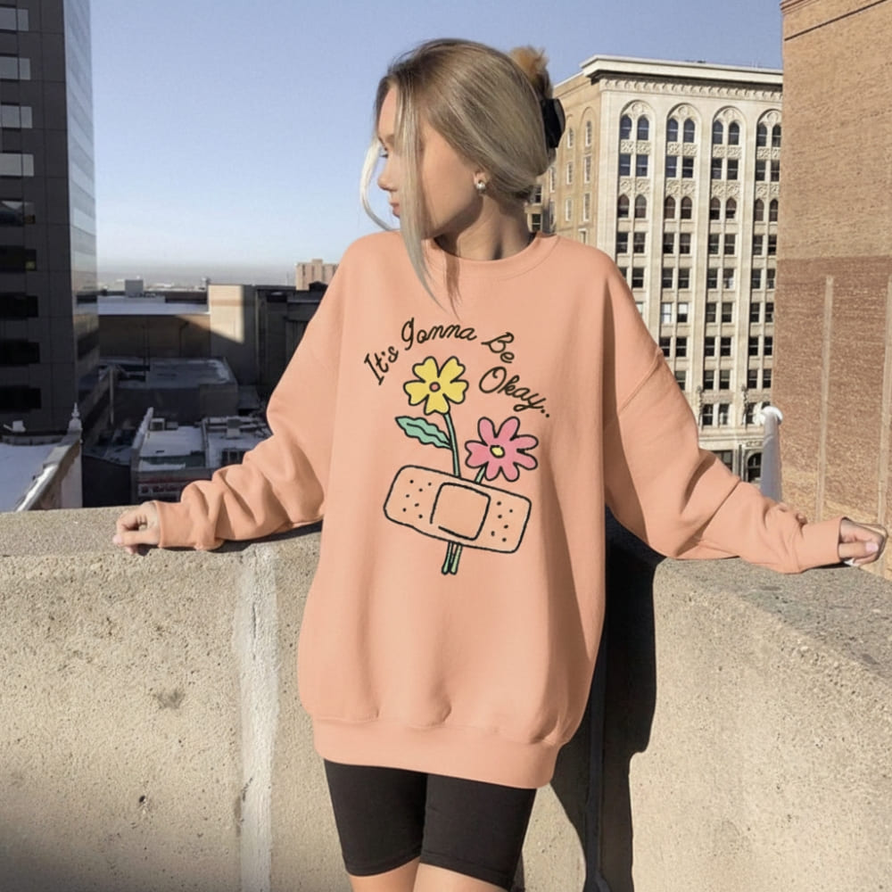 Person wearing a peach-colored sweatshirt with a graphic design on a rooftop with cityscape background