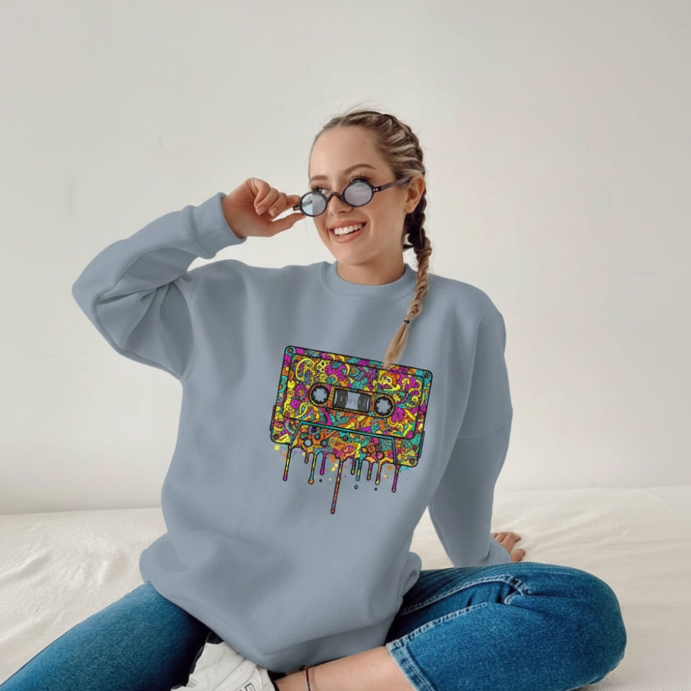 Person wearing a light blue sweatshirt with a colorful cassette tape design, sitting on a white surface.