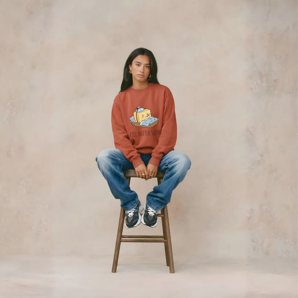 Person sitting on a stool wearing a red sweatshirt with a graphic design and blue jeans against a plain background