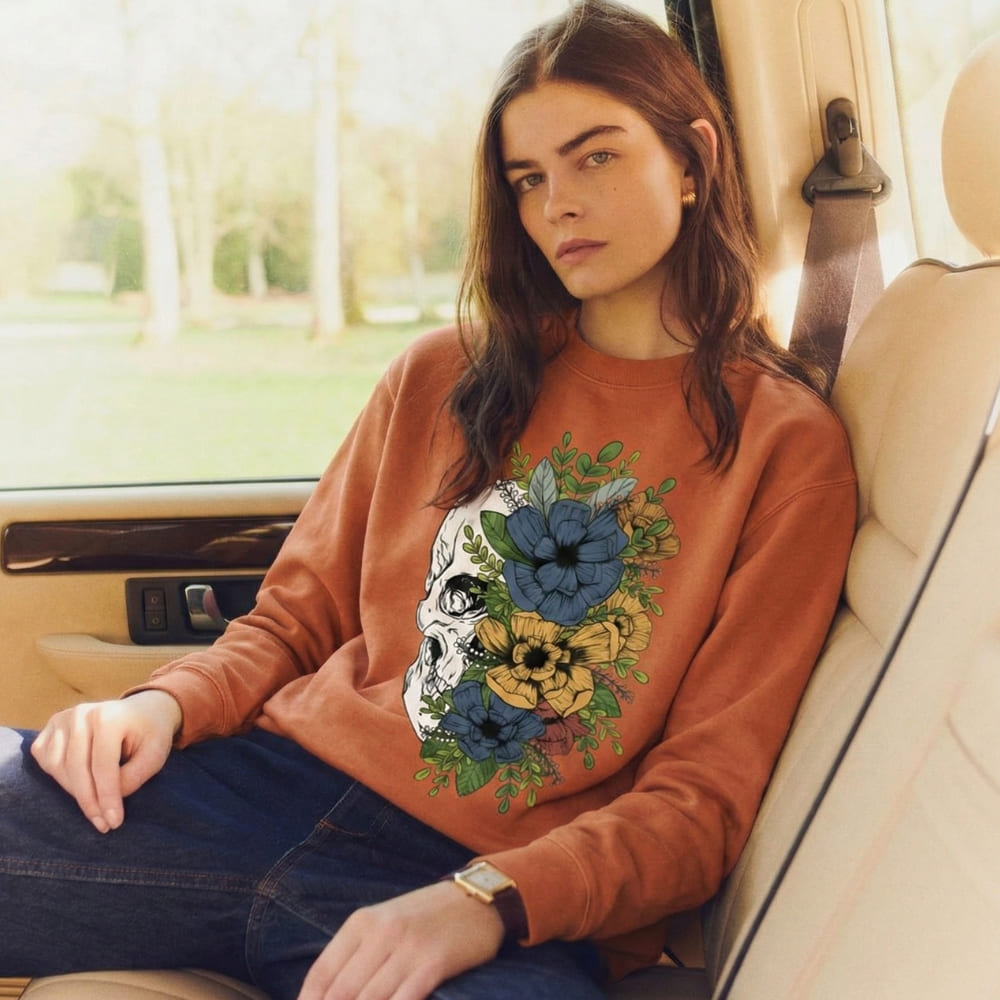 Woman wearing a orange sweatshirt with a floral and skull design, sitting inside a car.