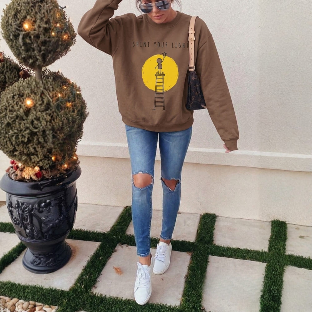 Person wearing a brown sweatshirt with a graphic design, blue jeans, and white sneakers standing next to a decorative plant.