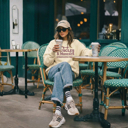 Person sitting at an outdoor cafe table holding a coffee cup, wearing a sweatshirt with text.