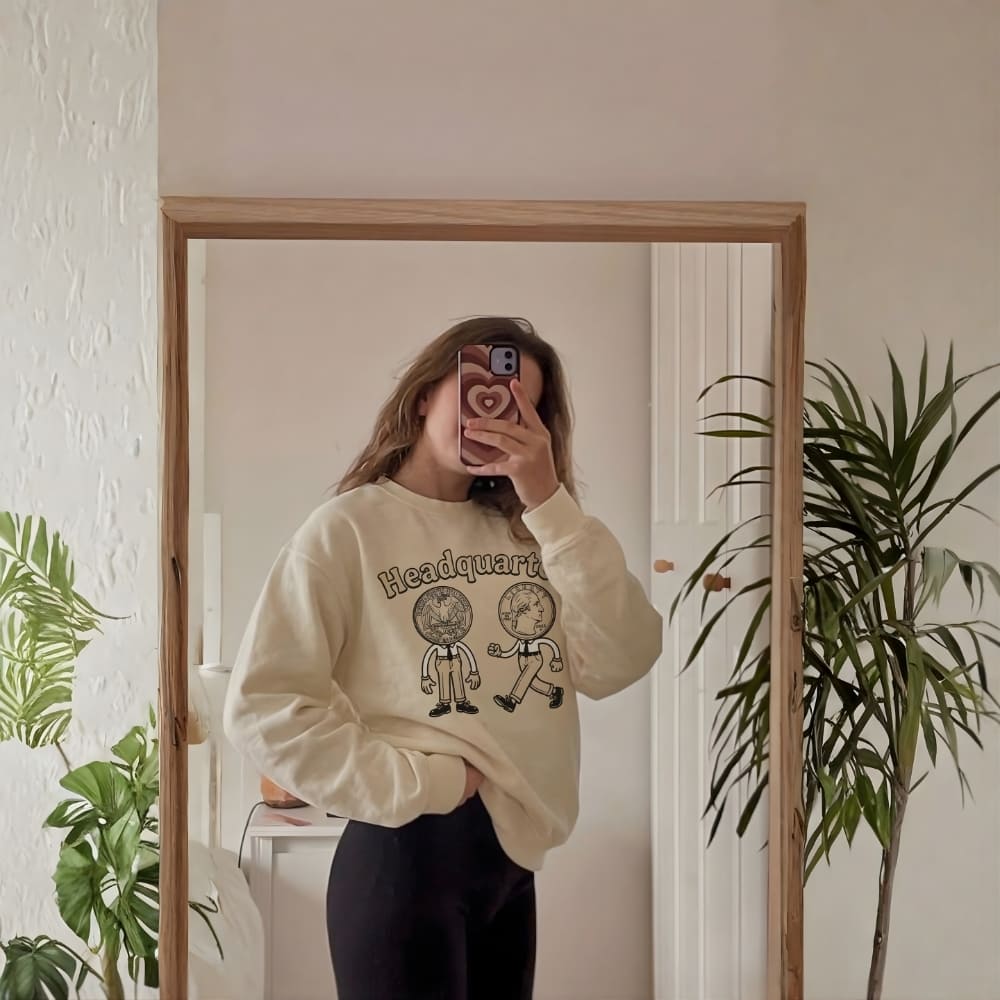 Person wearing a cream-colored sweatshirt with graphics in front of a mirror, surrounded by indoor plants.