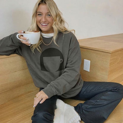 Woman sitting on a wooden floor holding a white mug, wearing a gray sweatshirt with a black graphic design.