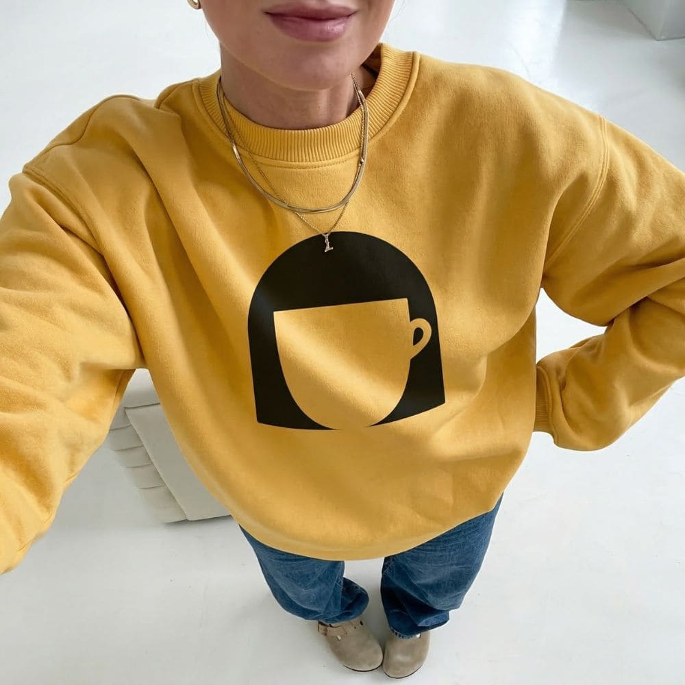 Person wearing a mustard yellow sweatshirt with a black graphic design on a white background
