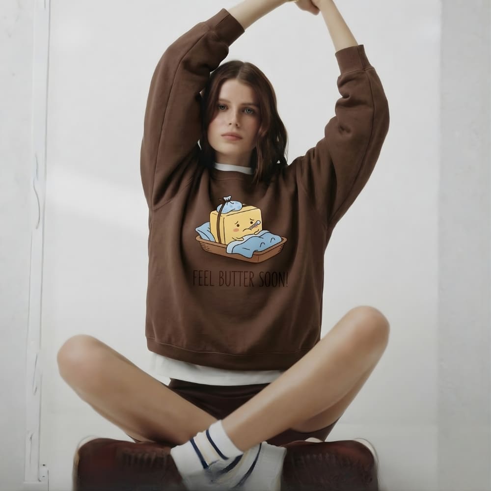 Person wearing a brown sweatshirt with a graphic design sitting on a white floor.