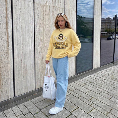 Person wearing a yellow sweatshirt with text, light blue jeans, and white sneakers, holding a Nike bag, standing on a sidewalk.