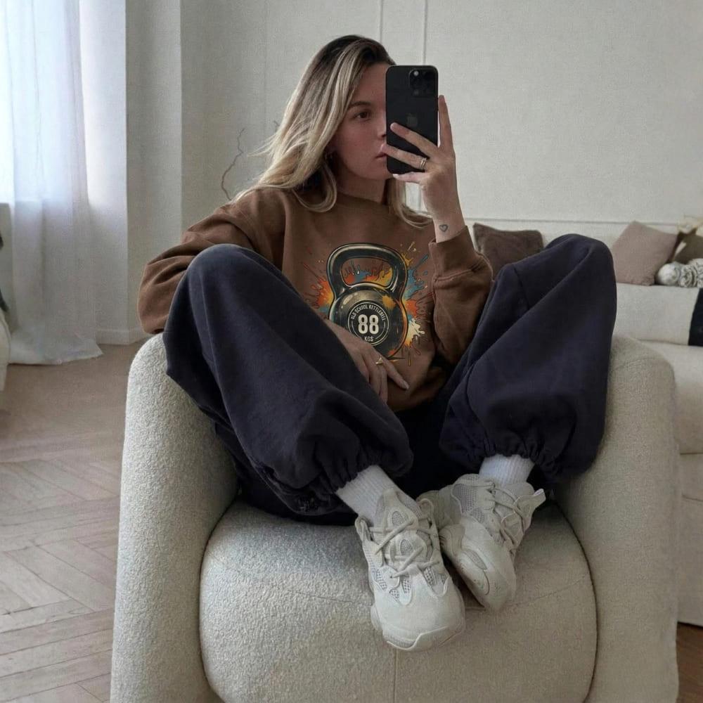 Person sitting on a couch wearing a brown sweatshirt with a graphic design, dark pants, and white sneakers.