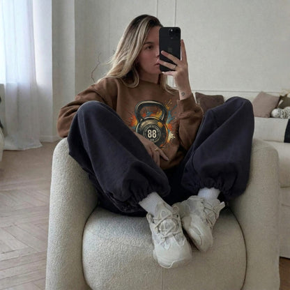 Person sitting on a couch wearing a brown sweatshirt with a graphic design, dark pants, and white sneakers.
