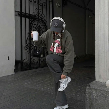 Person wearing a gray sweatshirt with a graphic design, gray pants, and white sneakers, holding a coffee cup and headphones.