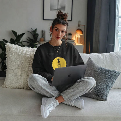Woman sitting on a couch with a laptop, wearing a black sweatshirt with a yellow design and gray pants.