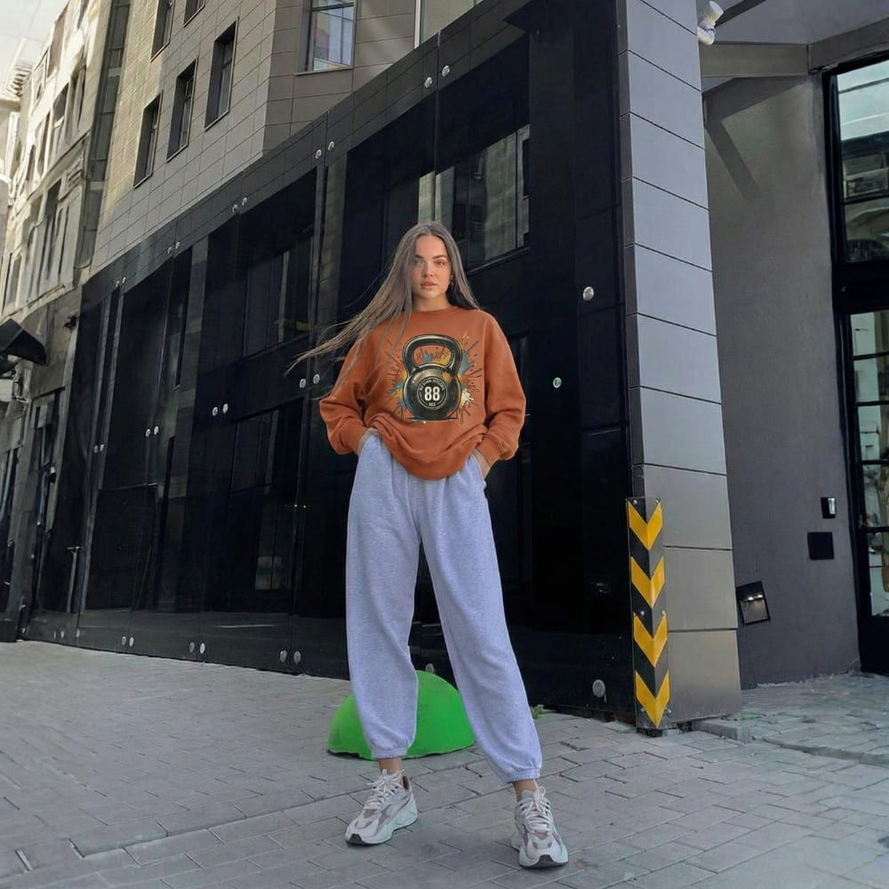 Woman wearing a brown sweatshirt with a kettlebell design and light gray sweatpants standing on a city street.