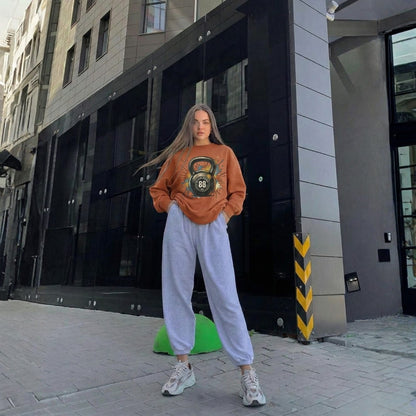 Woman wearing a brown sweatshirt with a kettlebell design and light gray sweatpants standing on a city street.