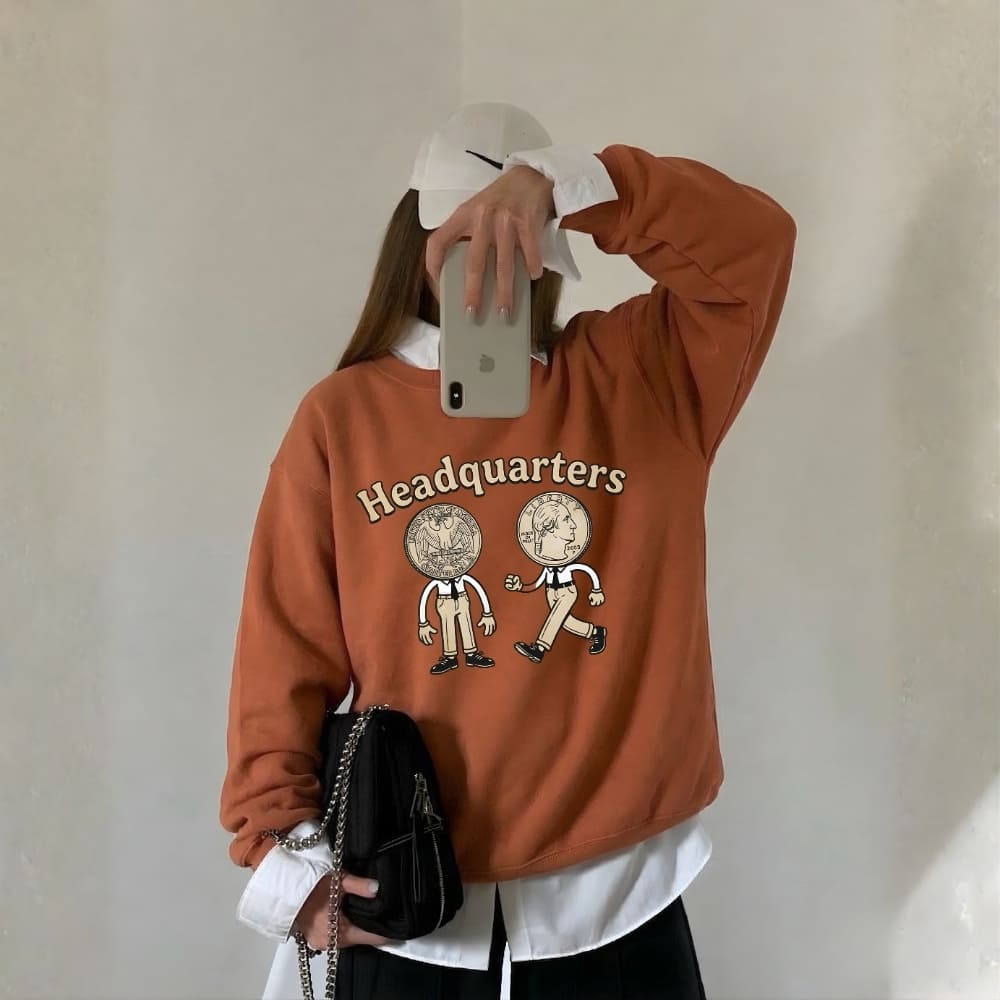 Person wearing a orange sweatshirt with 'Headquarters' and cartoon designs, holding a phone.