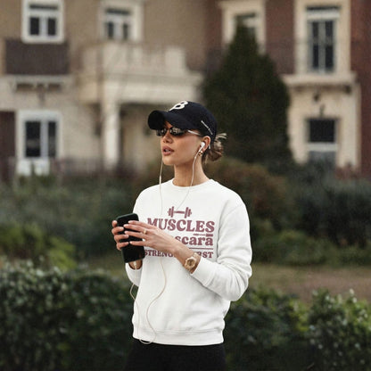 Woman wearing a white sweatshirt with text, black cap, and sunglasses, holding a phone outdoors.