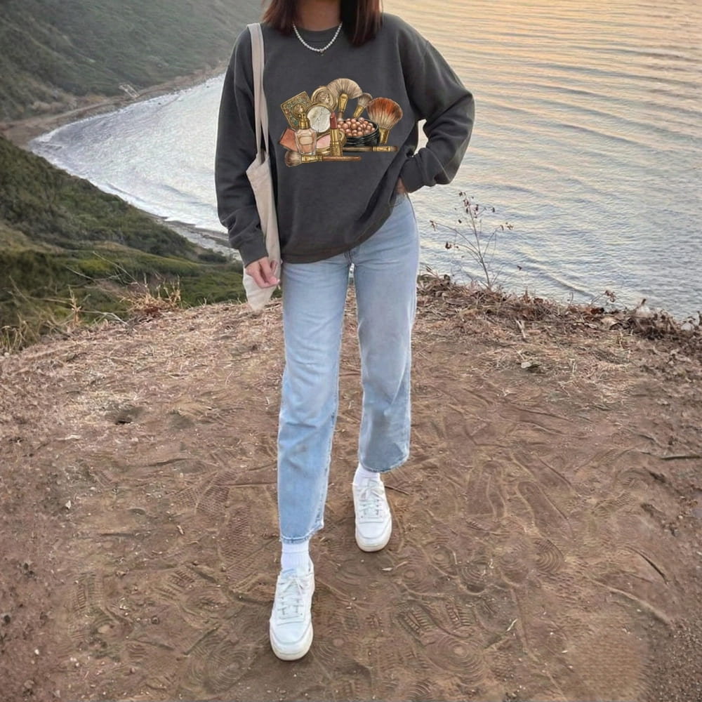 Person wearing a dark gray sweatshirt with a graphic design, light blue jeans, and white sneakers standing on a dirt path with a scenic background.