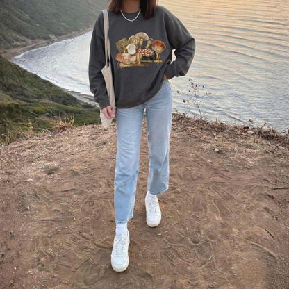 Person wearing a dark gray sweatshirt with a graphic design, light blue jeans, and white sneakers standing on a dirt path with a scenic background.