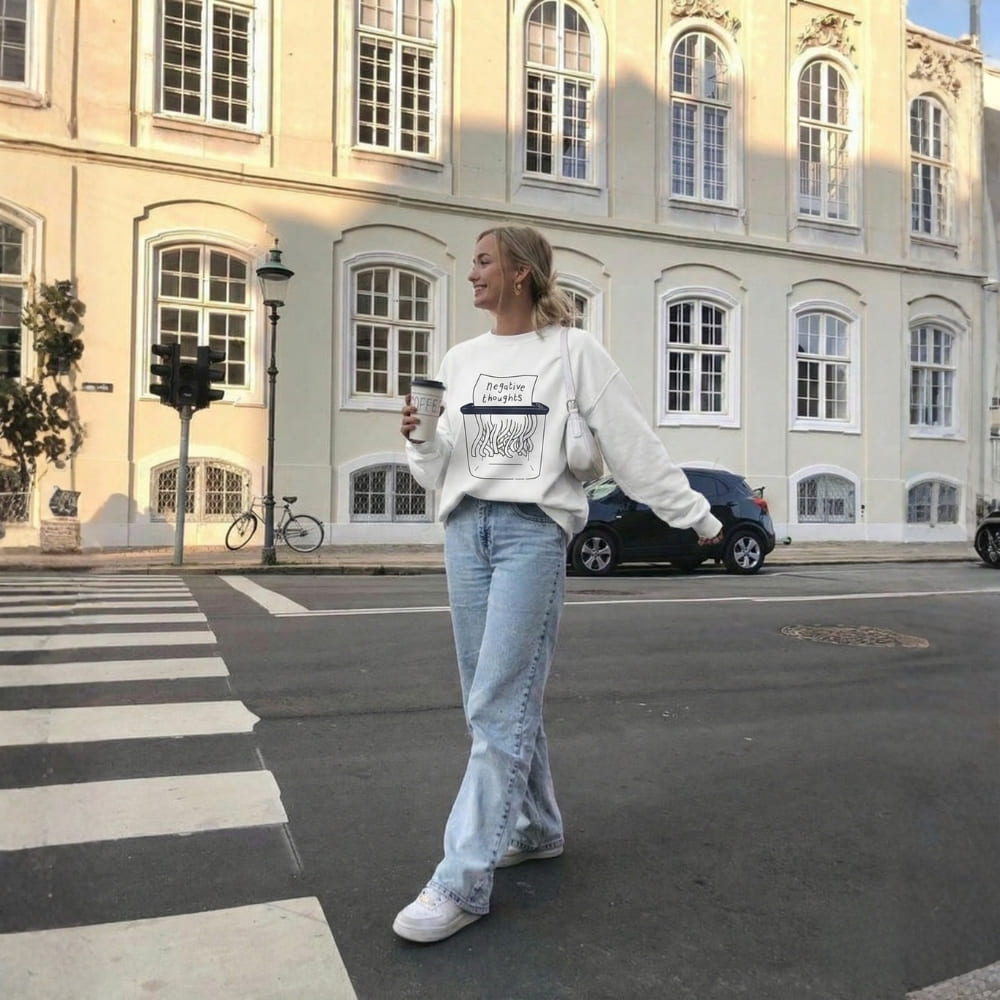 Person wearing a white sweatshirt with text and light blue jeans, standing on a street with a building in the background.