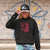 Person wearing a black hoodie with a graphic design and a cap, standing against a graffiti-covered wall.