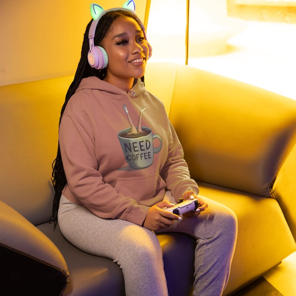 Woman sitting on a couch wearing a hoodie with 'Need Coffee' text, headphones, and holding a phone.