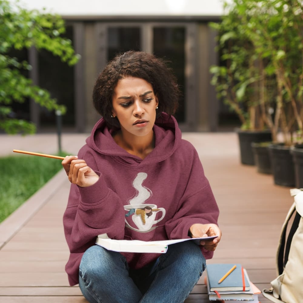Woman sitting outdoors holding a pencil and book, wearing a maroon hoodie with a graphic design.