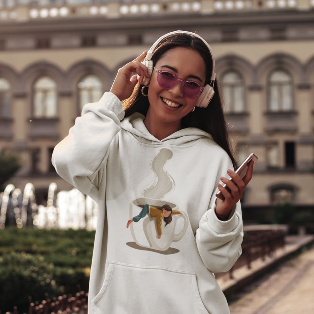 Woman wearing a white hoodie with a graphic design, pink headphones, and holding a phone outdoors.