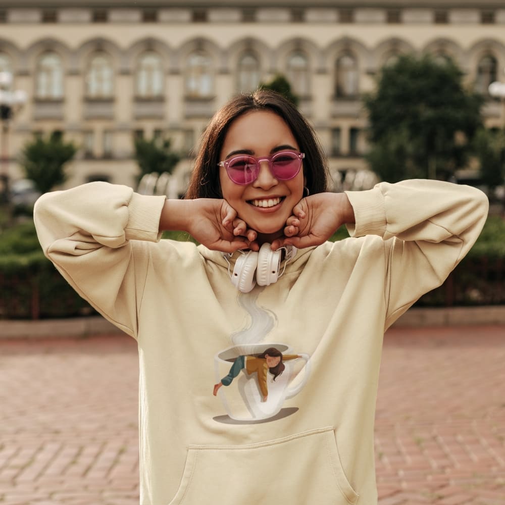 Person wearing a beige hoodie with a graphic design, standing outdoors with a blurred building in the background.