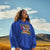 Person wearing a blue hoodie with a graphic design, standing in a scenic outdoor setting with mountains and blue sky.