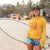 Woman in yellow hoodie and colorful shorts holding a surfboard on a beach.