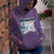Person wearing a purple hoodie with a graphic design, standing against a brick wall.