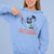 Person wearing a light blue hoodie with a graphic design and text, pointing to the design.